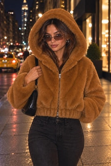 Styling image of a model wearing a copper hooded faux fur zip-front jacket with black jeans on a city street at night.